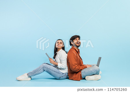 Sitting back to back, indian couple engages with technology separately, a man with a laptop and woman with a tablet on a blue backdrop Sitting back to back, indian couple engages with technology separately, a man with a laptop and woman with a tablet on a blue backdrop 114705260