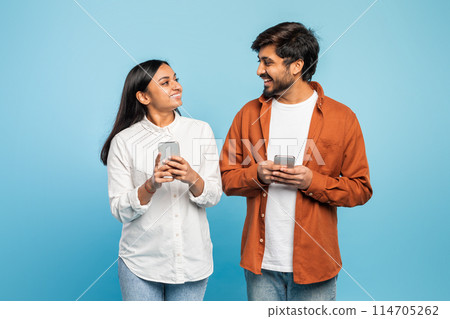 A happy indian couple holding phones and looking at each other against a blue background 114705262