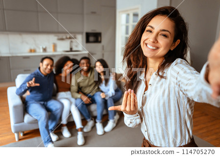 A cheerful young woman takes a selfie with her diverse group of friends chilling on the couch in the background 114705270