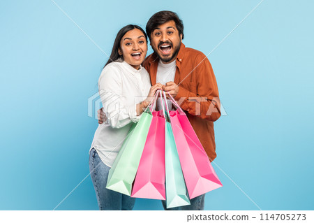 An ecstatic indian couple holds shopping bags up high, expressing excitement with wide smiles on a blue backdrop 114705273