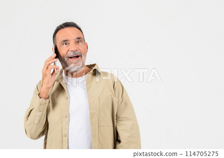 A cheerful elderly man with gray beard is engaging in a conversation on his mobile phone, expressing happiness and surprise 114705275