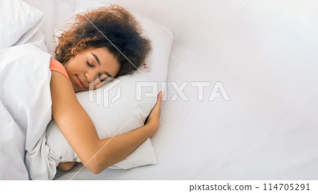 Relaxed african-american woman sleeping on bed and hugging pillow at home Relaxed african-american woman sleeping on bed and hugging pillow at home 114705291