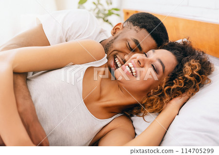 Happy millennial black couple flirting in morning on comfortable bed, tickling and laughing Happy millennial black couple flirting in morning on comfortable bed, tickling and laughing 114705299