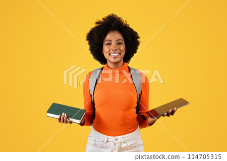 Smiling young African American woman student with a backpack holding a closed book in one hand and an open digital tablet in the other, against a yellow background, studio 114705315