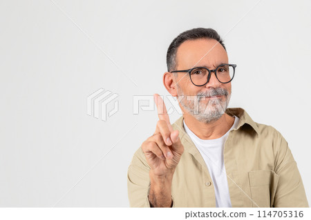 A confident elderly man with glasses pointing upwards with one finger and smiling, set against a white background 114705316