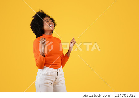 Joyful glad African American woman winner with closed eyes and a wide smile, dancing and enjoying the moment in an orange turtleneck on a yellow background, studio. Win, success 114705322