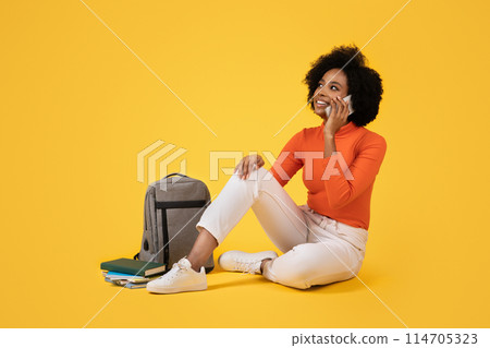 Thoughtful young woman sitting cross-legged, talking on the phone, dressed in an orange turtleneck, white pants, with books and a grey backpack beside her on a yellow background Thoughtful young woman sitting cross-legged, talking on the phone, dressed in an orange turtleneck, white pants, with books and a grey backpack beside her on a yellow background 114705323