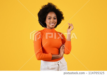 Confident smiling African American woman with curly hair poses in a stylish orange turtleneck, gracefully gesturing with one hand, against a vibrant yellow background, studio 114705325