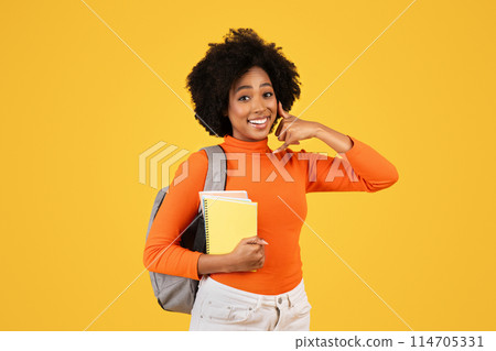 Cheerful young woman with curly hair holding notebooks, making a call me gesture, clad in an orange turtleneck and white pants, with a grey backpack on a sunny yellow background Cheerful young woman with curly hair holding notebooks, making a call me gesture, clad in an orange turtleneck and white pants, with a grey backpack on a sunny yellow background 114705331
