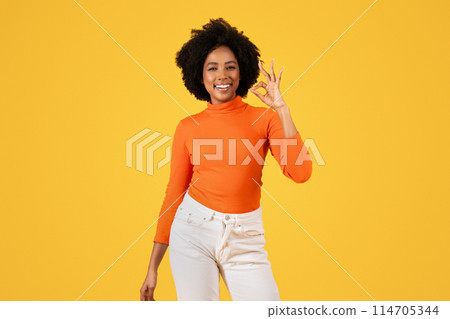 Contented woman with curly hair in orange turtleneck and white pants makes the OK sign, flashing a radiant smile, with a vibrant yellow backdrop highlighting her cheerful demeanor Contented woman with curly hair in orange turtleneck and white pants makes the OK sign, flashing a radiant smile, with a vibrant yellow backdrop highlighting her cheerful demeanor 114705344