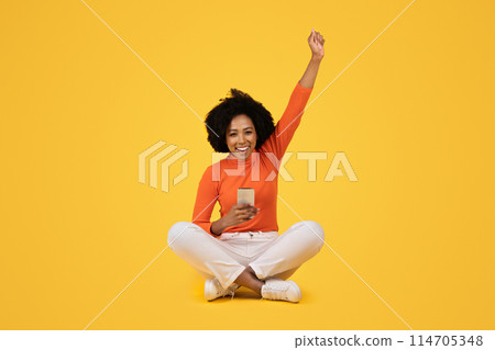 Joyful woman sitting cross-legged, celebrating with one arm raised, holding a smartphone in the other hand, dressed in an orange top and white pants against a yellow background 114705348