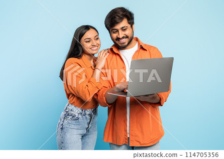 Indian couple in orange attire smiling while collaborating and looking at laptop screen, modern work concept 114705356