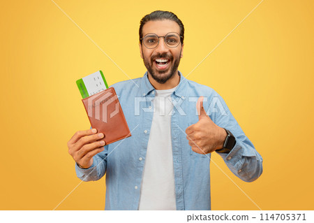 Joyful indian man shows approval with passport and boarding passes on yellow, signaling positive travel experience 114705371
