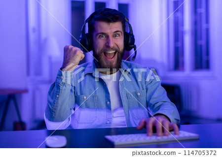 Excited guy gamer in headphones gestures a win while playing online games illuminated by computer screen 114705372