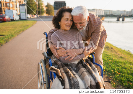Senior woman in wheelchair walking with caregiver old man on road in park. Elderly family couple man supporting embracing woman in chair for people with disability outdoor. Rehabilitation 114705373