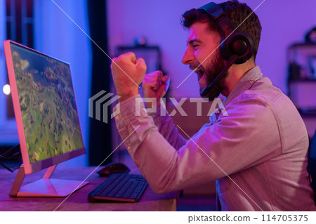 A man in a casual denim shirt is clenching his fist in frustration at a computer screen in a vibrant home office setting 114705375