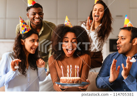 Multiracial friends surround a woman holding a birthday cake, blowing out candles in a moment of excitement together 114705381