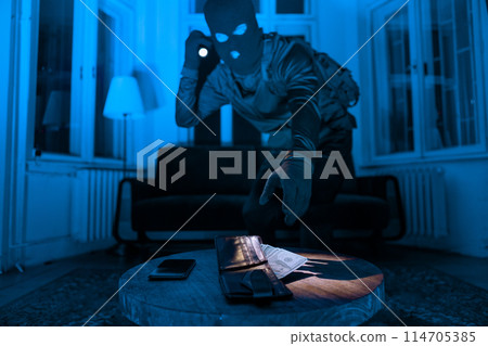 A masked burglar in a dark room uses a flashlight to find and steal a wallet and phone on a wooden table 114705385
