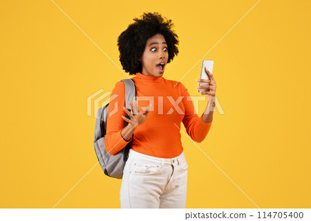 Surprised African American woman with curly hair, wearing an orange turtleneck and a grey backpack, reacts to content on her smartphone with a wide-eyed expression, on a yellow backdrop 114705400