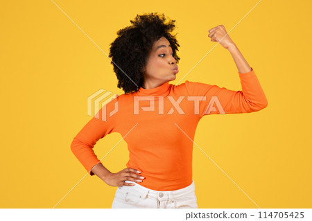 Confident young woman with curly hair flexing her arm muscle in a strong pose, wearing an orange turtleneck and white pants, with a playful pout, against a monochrome yellow background 114705425