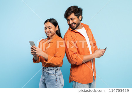 Casual young indian couple using their phones, engrossed in personal digital worlds with blue backdrop Casual young indian couple using their phones, engrossed in personal digital worlds with blue backdrop 114705429