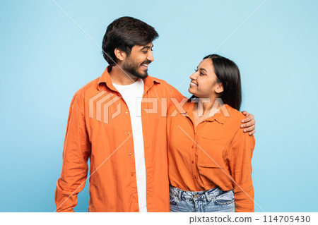 An indian man and woman stand close together in casual orange shirts, making a heartwarming display against a solid blue backdrop, faces blurred An indian man and woman stand close together in casual orange shirts, making a heartwarming display against a solid blue backdrop, faces blurred 114705430