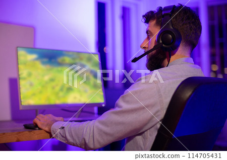 A man wearing a casual shirt with headphones intently uses a gaming computer in a room with colorful ambient lighting A man wearing a casual shirt with headphones intently uses a gaming computer in a room with colorful ambient lighting 114705431