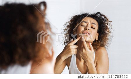 Afro girl frowning squeezing pimple on her face in mirror. Do not squeez pimples concept, copy space 114705432