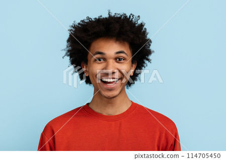 A joyful young black man with curly hair wearing a red sweater smiles against a blue background A joyful young black man with curly hair wearing a red sweater smiles against a blue background 114705450