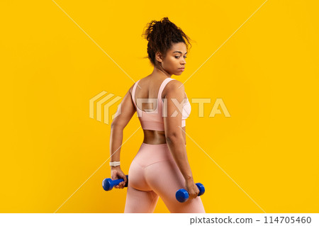 A strong african american woman confidently lifts blue dumbbells during her fitness session, showcasing strength on a yellow backdrop 114705460