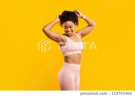 A vibrant young african american woman in pink workout attire happily prepares her hair for a fitness session against a yellow wall A vibrant young african american woman in pink workout attire happily prepares her hair for a fitness session against a yellow wall 114705468