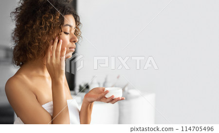 Black Young Woman Holding Face Cream Jar Standing In Bathroom. Skincare Routine. 114705496