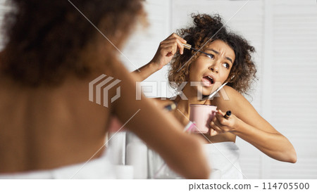 Afro-american girl in hurry put on makeup, drinking coffee and talking by phone simultaneously in front of mirror in bathroom. Morning of modern woman concept Afro-american girl in hurry put on makeup, drinking coffee and talking by phone simultaneously in front of mirror in bathroom. Morning of modern woman concept 114705500
