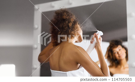 Young afro woman using thermal spray standing in front of mirror in bathroom. Young afro woman using thermal spray standing in front of mirror in bathroom. 114705503