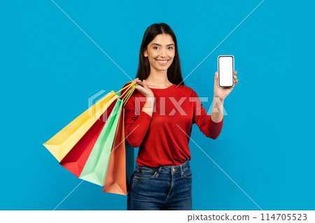 Woman holding multiple shopping bags and showing blank cell phone, happy female recommending new app for purchases, standing on blue studio background, mockup 114705523