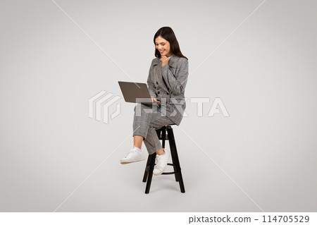 Relaxed and focused young businesswoman browsing on laptop while sitting on chair, displaying casual work style, against light background 114705529