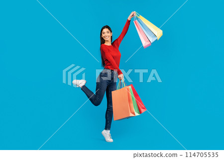 Excited young woman joyfully jumping in the air while holding shopping bags, showcasing her excitement for her purchases, posing on blue background 114705535