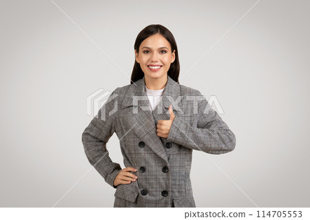 Radiant businesswoman giving thumbs up in approval, wearing checkered blazer, exuding confidence and positivity against neutral background, perfect for corporate and success themes 114705553