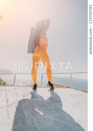 Fitness woman sea. Happy middle aged woman in orange sportswear exercises morning outdoors on yoga mat with laptop in park over ocean beach. Female fitness pilates yoga routine. Healthy lifestyle. 114705561