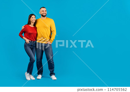 Smiling couple standing together, man with arm around woman, both looking to the side at copy space, happy spouses posing against vivid blue studio background, full length 114705562