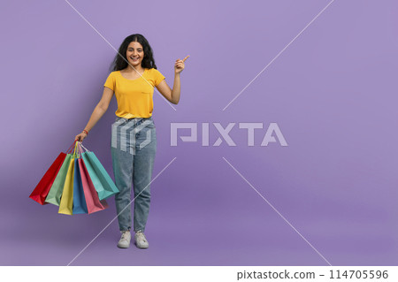 Cheerful woman holding shopping bags and pointing upwards with a smile on a purple background Cheerful woman holding shopping bags and pointing upwards with a smile on a purple background 114705596