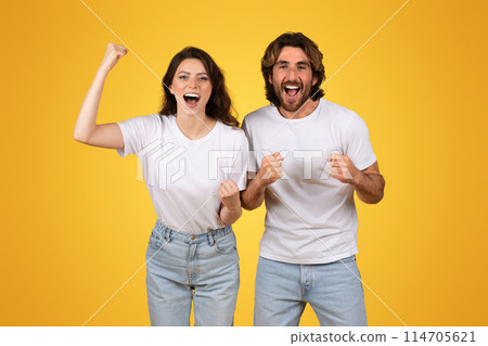 Energetic european young couple with raised fists celebrating a victory, exuding joy and excitement, wearing white t-shirts and jeans against a vivid yellow background, studio 114705621