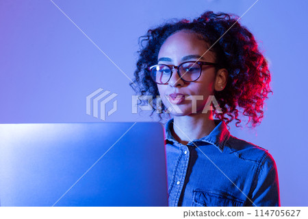 Professional young black woman with curly hair and glasses concentrating on her laptop, illuminated by soft blue light on dual-tone background Professional young black woman with curly hair and glasses concentrating on her laptop, illuminated by soft blue light on dual-tone background 114705627