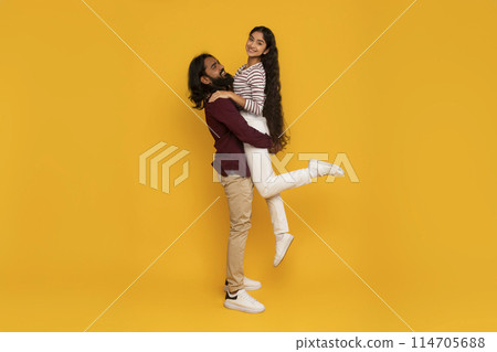 A happy man with a beard is smiling while carrying a woman in a joyful pose against a vivid yellow background A happy man with a beard is smiling while carrying a woman in a joyful pose against a vivid yellow background 114705688