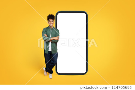 Confident african american man in casual clothes leaning on a gigantic smartphone mockup against a yellow backdrop Confident african american man in casual clothes leaning on a gigantic smartphone mockup against a yellow backdrop 114705695