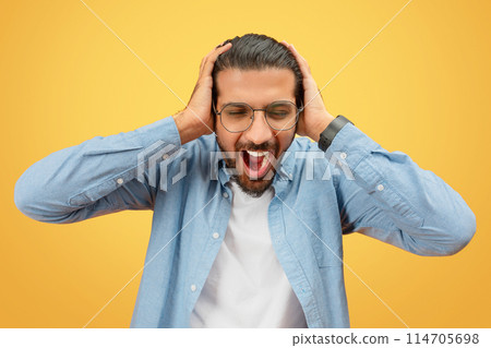 A distraught indian man holds his head and screams, displaying a sense of desperation on a yellow background 114705698