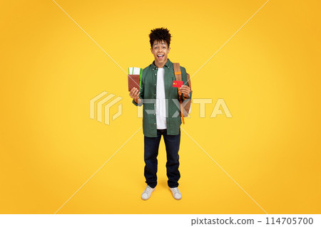 Smiling black guy with backpack holding a passport and a boarding pass ready for travel on a yellow background, using credit card, booking tickets 114705700