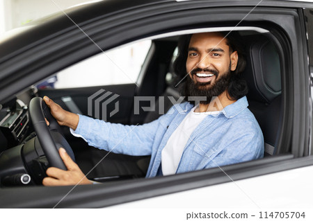 A cheerful bearded indian man with long hair is sitting behind the wheel of a contemporary vehicle, looking at the camera with a broad smile 114705704
