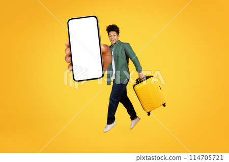 Cheerful african american guy holds out a smartphone screen and jumps with a suitcase on a yellow background, showing excitement and mobility 114705721