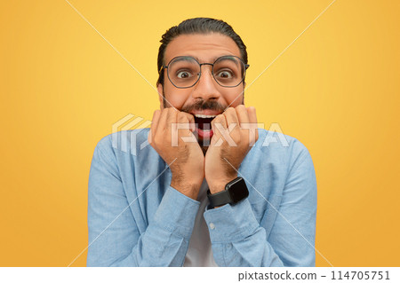 An anxious looking indian man with hands on his face against a plain yellow background, portraying stress An anxious looking indian man with hands on his face against a plain yellow background, portraying stress 114705751